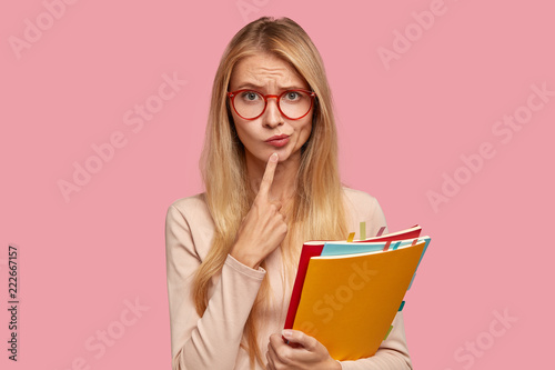 Puzzled schoolgirl has hesitant expression, purses lips, keeps index finger on chin, wears round spectacles, tries to decide what do after classes, carries books, isolated over pink background