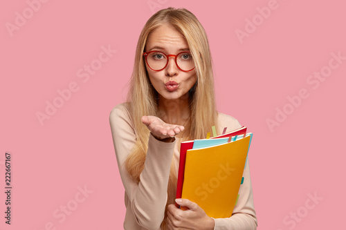 Horizontal shot of pleasant looking woman with blonde hair, blows air kiss to boyfriend, wears spectacles, studies at college or university, carries textbook, isolated over pink studio wall.