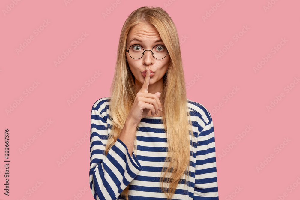 Studio shot of secret young woman keeps fore finger over lips, has blonde hair, makes shush gesture, dressed in striped sailor sweater, stands over pink background. People and conspiracy concept Studio shot of secret young woman keeps fore finger over lips, has blonde hair, makes shush gesture, dressed in striped sailor sweater, stands over pink background. People and conspiracy concept