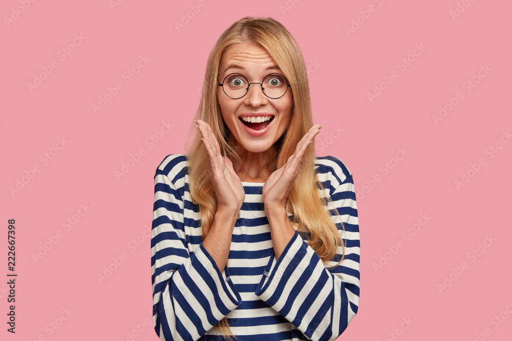 Excited happy impressed girl clasps hands near face, notices something thrilling, dressed in casual striped sweater, expresses positive emotions, stands alone against pink background. Wow, its cool! Excited happy impressed girl clasps hands near face, notices something thrilling, dressed in casual striped sweater, expresses positive emotions, stands alone against pink background. Wow, its cool!