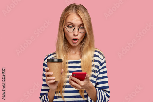 Isolated shot of surprised blonde girlfriend shocked to recieve message from lover about his attention to part, stares with terrible gaze at cell phone, carries disposable coffee, wears sailor sweater