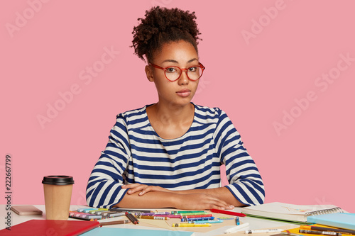 Pretty serious dark skinned woman likes painiting, spends free time drawing picture on blank sheet, uses crayons, wears transparent glasses, sailot sweater, drinks takeaway coffee, sits indoor photo