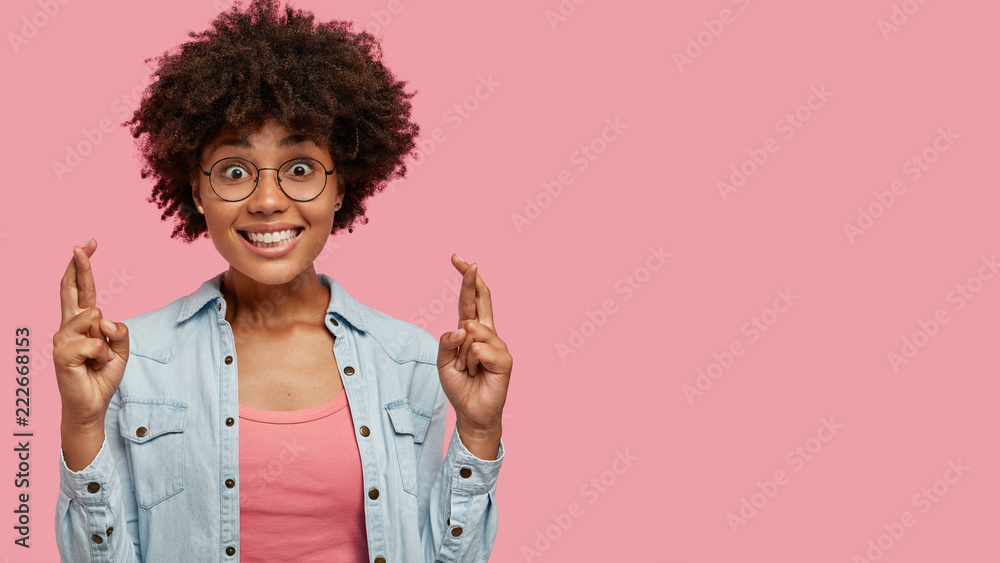 Horizontal shot of cheerful girlfriend with broad shing smile, keeps fingers crossed, believes in good luck and wellness, prays indoor, stands against pink background with copy space for your text Horizontal shot of cheerful girlfriend with broad shing smile, keeps fingers crossed, believes in good luck and wellness, prays indoor, stands against pink background with copy space for your text