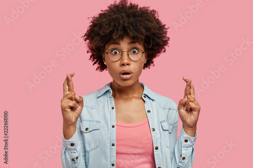 Horizontal shot of terrified dark skinned youngster crosses fingers for good luck, being stupefied with something, afraids of passing exam, dressed in denim jacket, isolated over pink background