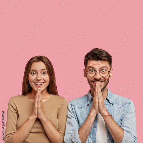 Isolated shot of cheerful pleased boyfriend and girlfriend keep palms in praying gesture, ask their dreams come true, stand next to each other, believe in something positive, isolate on pink wall