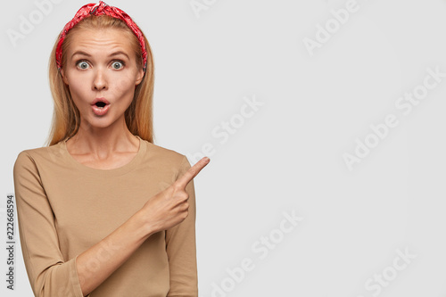 Image of surprised Caucasian woman with stupefied expression, points at upper right corner, shows blank space for your advertising content, dressed casually, stunned by something, has bated breath