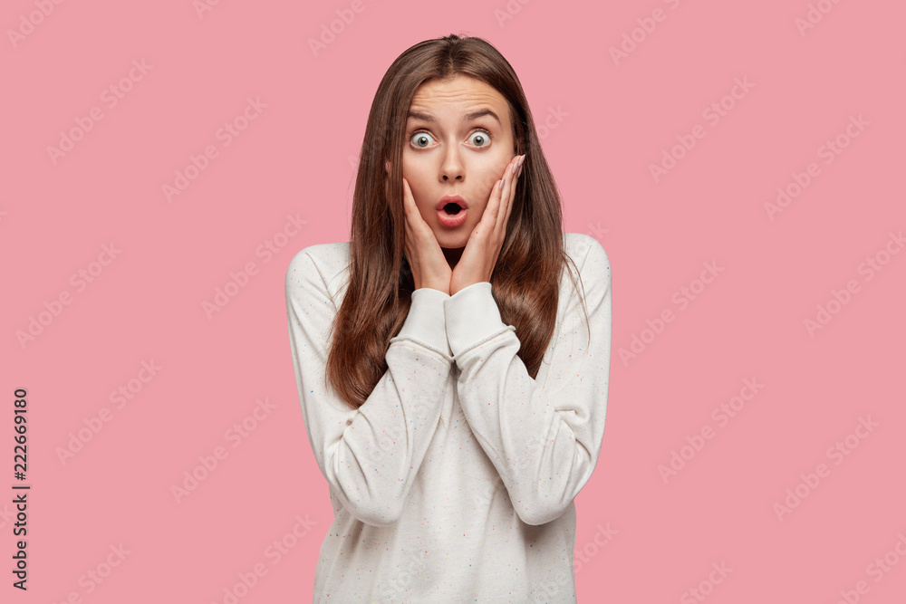Portrait of surprised brunette teenage girl, keeps mouth wide opened, wonders sudden news, dressed in loose sweatshirt, stands against pink studio wall. People, emotions and excitement concept. Portrait of surprised brunette teenage girl, keeps mouth wide opened, wonders sudden news, dressed in loose sweatshirt, stands against pink studio wall. People, emotions and excitement concept.