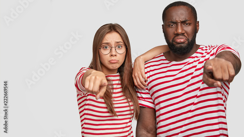 Isolated shot of dissatisfied woman and man of different nationalities, frown face in discontent, points directly at camera, dressed in casual striped t shirt, isolated over white background