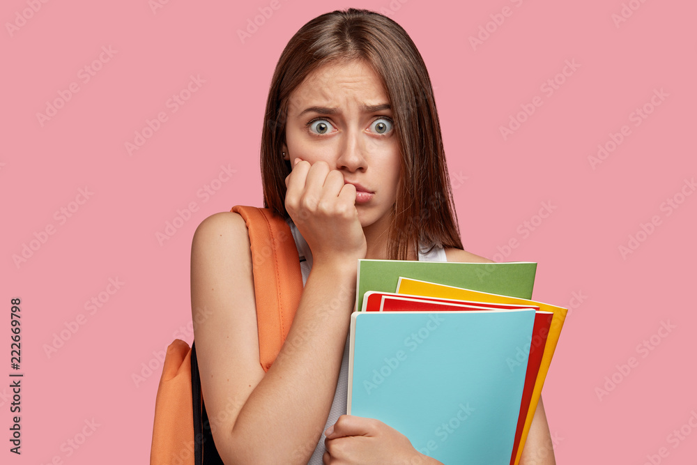 Troubled frightened schoolgirl stares with bugged eyes at camera, keeps hand near mouth, carries rucksack with necessary things, isolated over pink background. People, reaction and shock concept Troubled frightened schoolgirl stares with bugged eyes at camera, keeps hand near mouth, carries rucksack with necessary things, isolated over pink background. People, reaction and shock concept