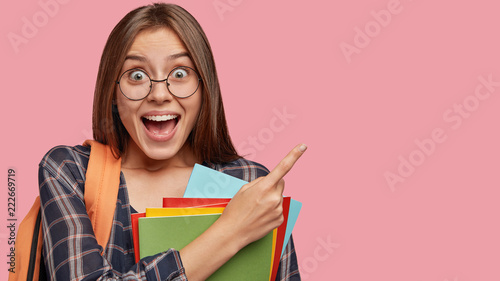 Attractive happy girl being in high spirit after classes, opens mouth and exclaims from happiness, points with index finger at copy space for your promotion, dressed in casual outfit, carries rucksack