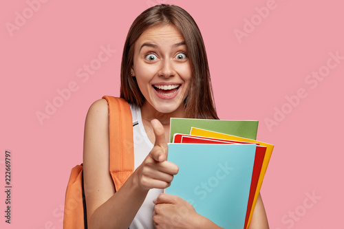 I choose you to help! Happy young Caucasian girl with pleasant appearance indicates directly at camera with index finger, dressed in casual clothes, carries rucksack, isolated over pink background.