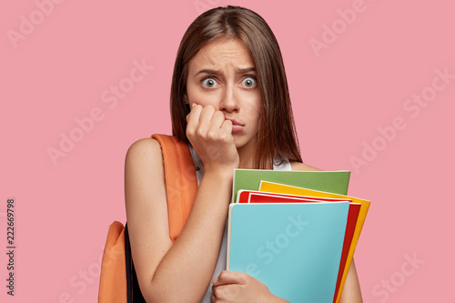 Troubled frightened schoolgirl stares with bugged eyes at camera, keeps hand near mouth, carries rucksack with necessary things, isolated over pink background. People, reaction and shock concept