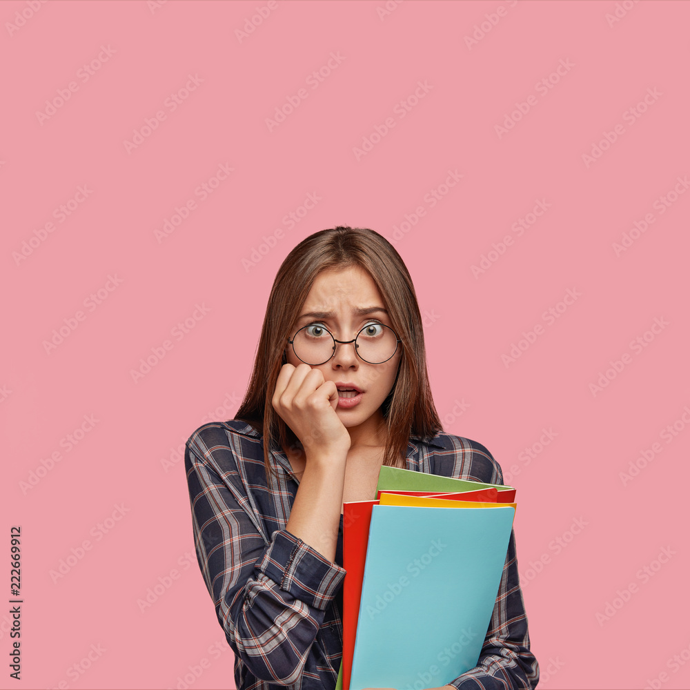Vertical shot of stupefied European woman keeps hand near mouth, looks with terror at camera, dressed in casul checkered shirt, finds out results of final exam, isolated over pink background. Vertical shot of stupefied European woman keeps hand near mouth, looks with terror at camera, dressed in casul checkered shirt, finds out results of final exam, isolated over pink background.