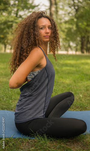 A girl is sitting in the park, practicing yoga, looking at the camera, curly hair. Summer day