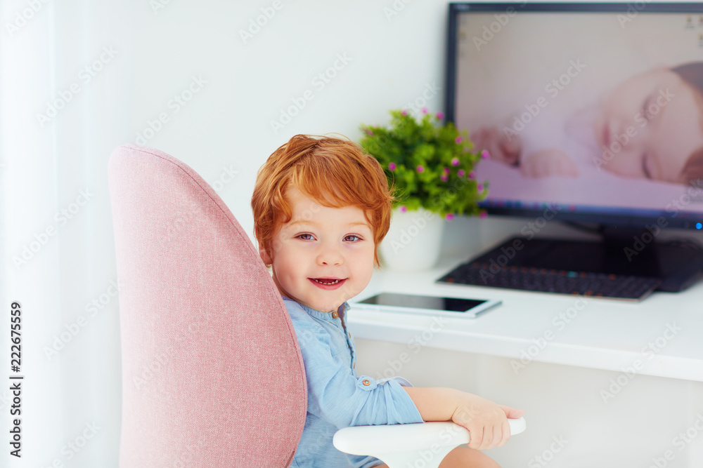 happy redhead toddler baby boy is sitting in office chair at working place