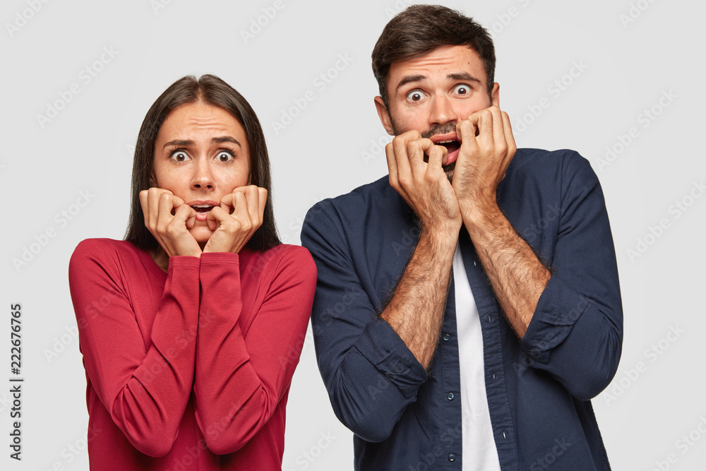 Φωτογραφία Fearful stunned emotional young woman and man bite fingers nervously, stare with frightened expression, feel anxious, notice phobia, dressed in casual outfit, stand against white background
