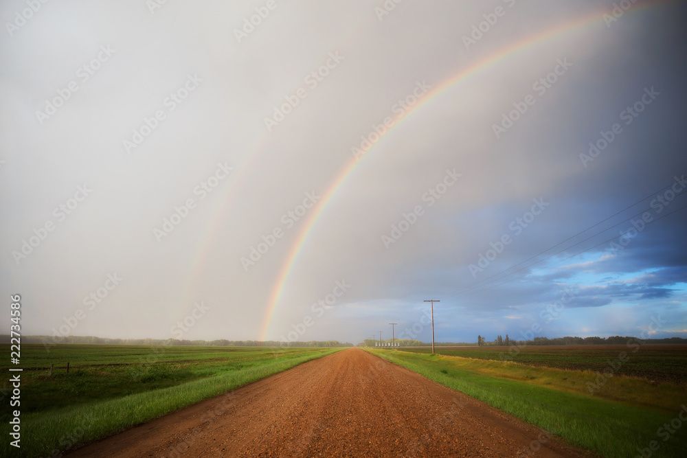 Half of a rainbow arching over a gravel road divivding green agricultural crops with a distant forest in a summer countryside landscape