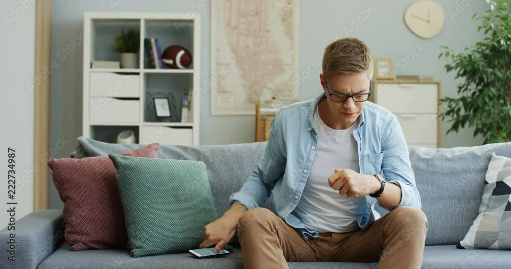 Young Caucasian man sitting on the couch and chatting on the phone with sms, then being upset and looking at the side. At home. Young Caucasian man sitting on the couch and chatting on the phone with sms, then being upset and looking at the side. At home.