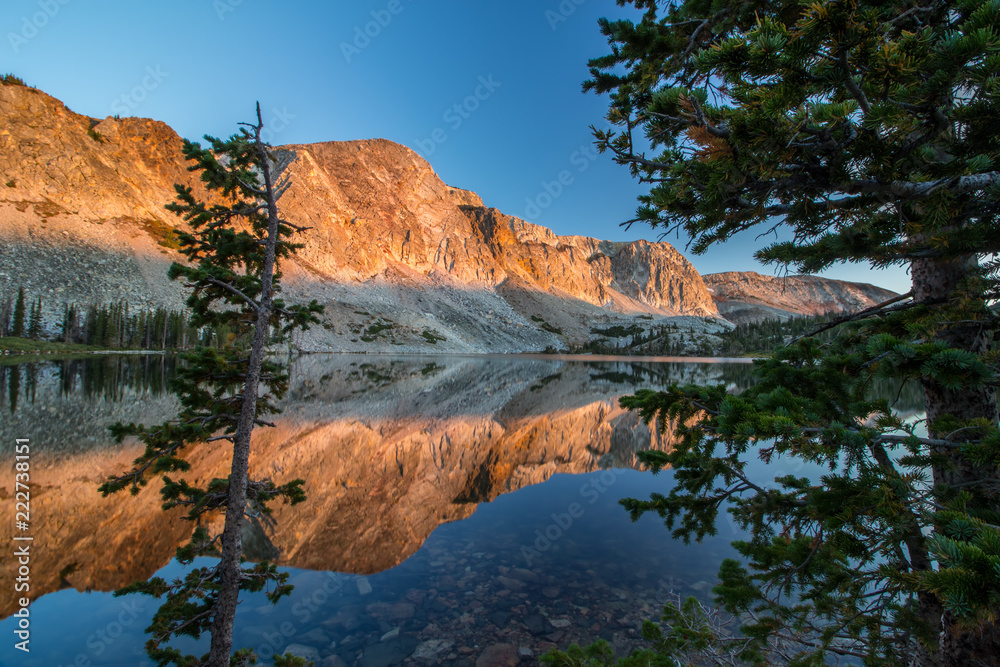 The light of sunrise strikes the sheer rock walls of the Snowy Range in Wyoming, reflecting off the still surface of a lake.