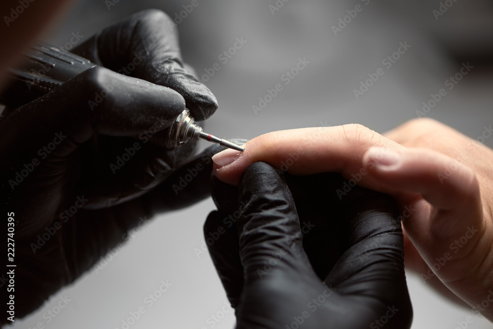 Close-up of professional master manicurist hands in black gloves working with electric drill file on client woman fingernails. Finger nail treatment, fashion, manicure making in beauty salon.
