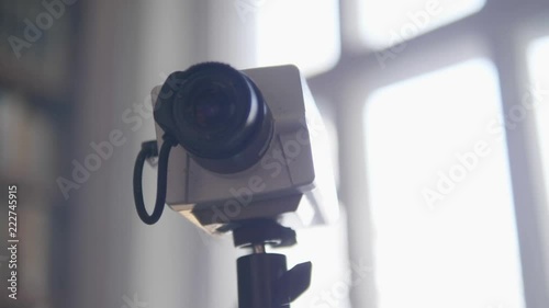 Demonstraion of old grey square security camera with small black cylinder shaped lens mounted to tripod in daylight room on blurry bright background. photo