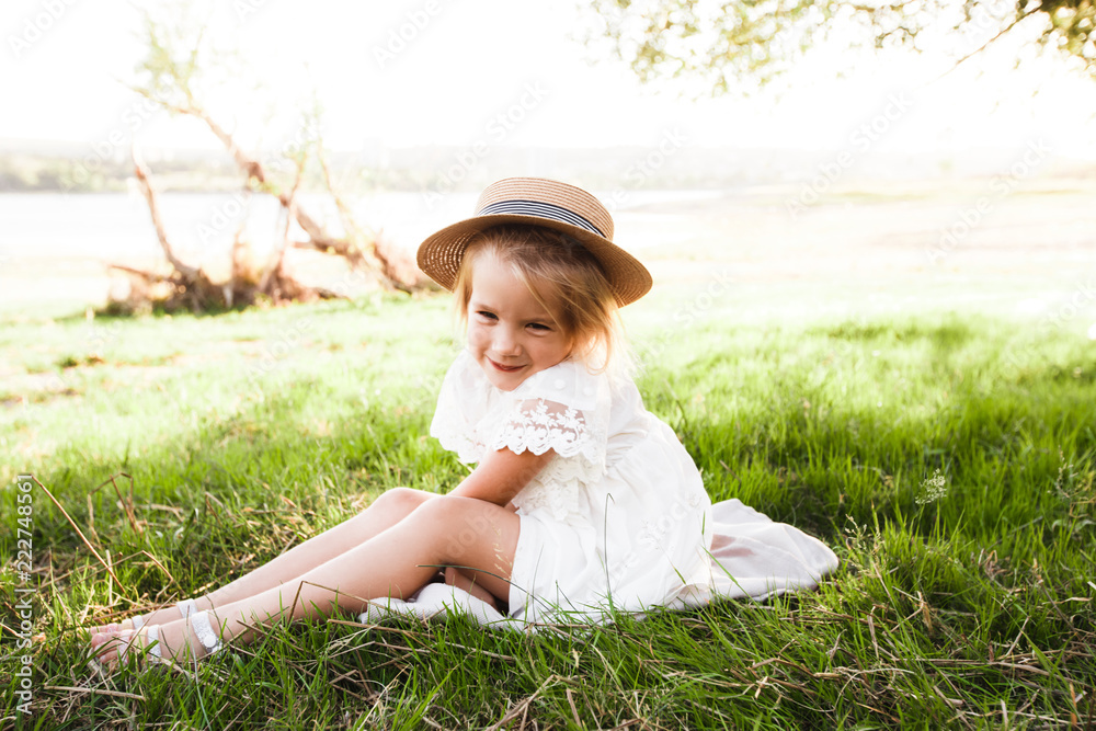 A cute baby girl in a straw hat and white dress is sitting on the grass. A cute baby girl in a straw hat and white dress is sitting on the grass.