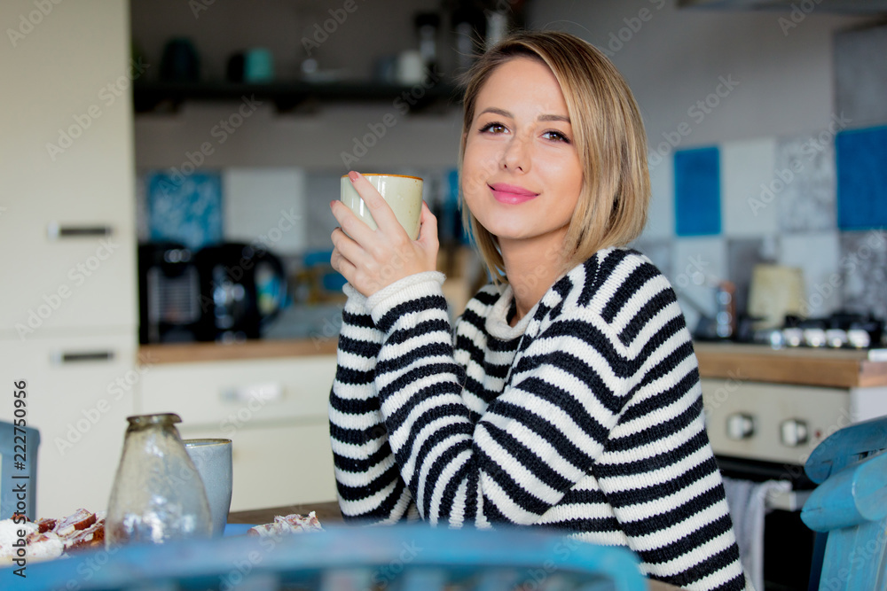 Young lady in sweater sitting at the table in breakfast time with cup of coffee and cake at the kitchen. - obrazy, fototapety, plakaty 