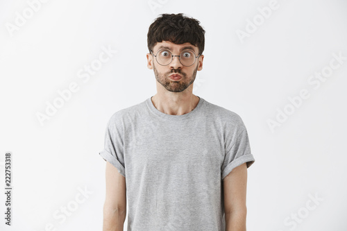 Waist-up shot of playful good-looking and stylish caucasian male model with beard in grey t-shirt holding breat and pouting popping eyes at camera making bet not to breath over gray wall