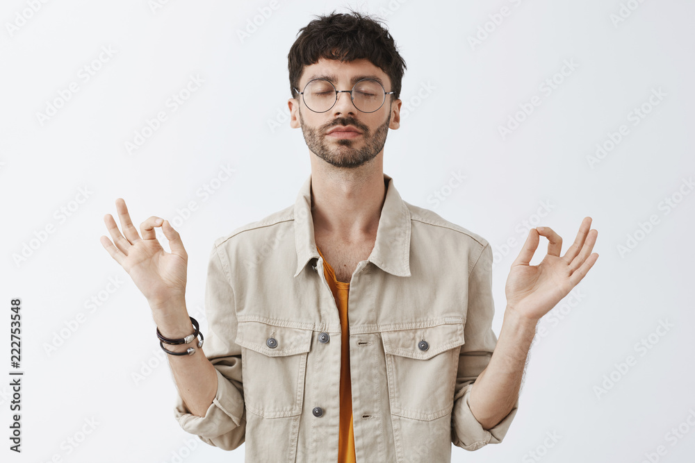 Calm good-looking and stylish urban guy with beard and trendy hairstyle in glasses meditating to relieve from stress being patient and relaxed practicing yoga with hands spread aside and closed eyes Calm good-looking and stylish urban guy with beard and trendy hairstyle in glasses meditating to relieve from stress being patient and relaxed practicing yoga with hands spread aside and closed eyes