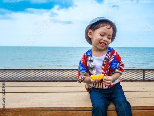 Cute happy asian boy in a summer shirt eating yellow lemon in hand on the wooden table posing background of Hua Hin sea blue skyThailand.Copy-space for your edit. photo