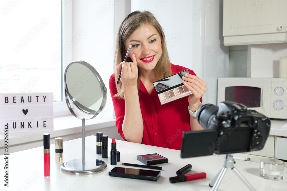 Closeup picture of young positive European female sitting at table with mirror and camera presenting nude eyeshadow palette to viewers pointing to some hues with brush to share her opinion about them Closeup picture of young positive European female sitting at table with mirror and camera presenting nude eyeshadow palette to viewers pointing to some hues with brush to share her opinion about them