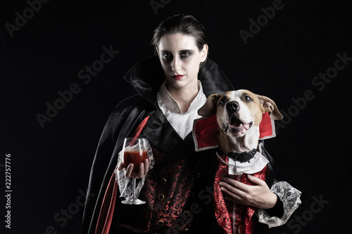 Woman and her dog in similar vampire costumes for halloween. Young female with glass of red drink and her pet puppy dressed up in same dracula costume.