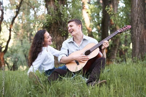 young couple sitting in the forest and playing guitar, summer nature, bright sunlight, shadows and green leaves, romantic feelings