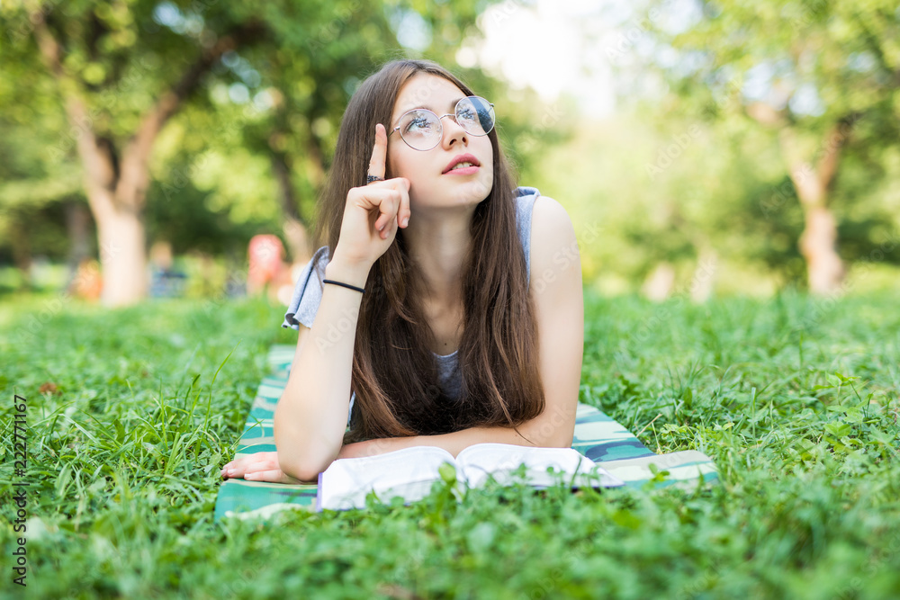 Concentrated young woman resting with book in park. Serious beautiful girl lying on grass while reading favorite romance. Dreaming with literature concept Concentrated young woman resting with book in park. Serious beautiful girl lying on grass while reading favorite romance. Dreaming with literature concept