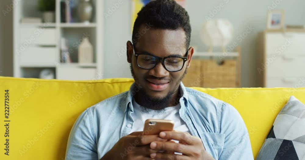 Portrait shot of the cheerful young African American man taping while texting on the smartphone device in the living room.