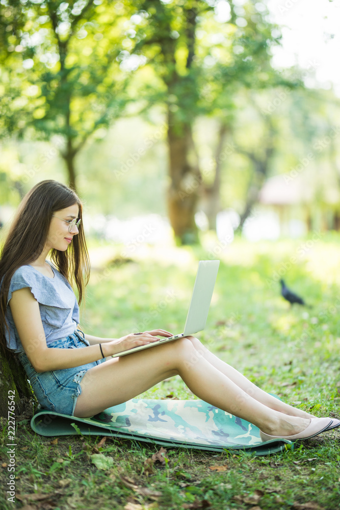 Young beautiful woman is sitting on green grass under the tree in the garden on summer day and working on her laptop
