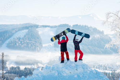 Two people stand with their backs on a snowdrift and raise their snowboards up against a white haze of snow-capped mountains and forests at winter ski resort.