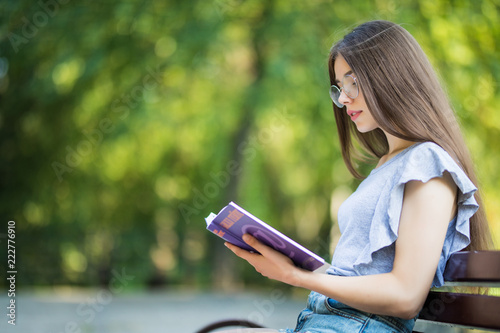 Side view of pleased woman in eyeglasses sitting on bench and reading book in park