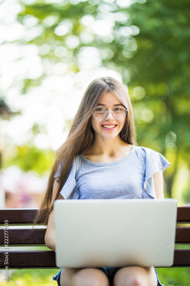 Young woman in eyeglasses sitting on bench in park and using laptop computer Young woman in eyeglasses sitting on bench in park and using laptop computer