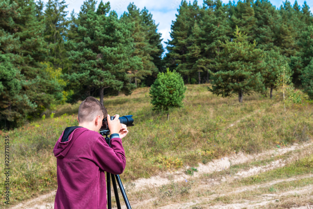 Young unidentifiable teenage boy taking landscape images with a camera on tripod near a pine forest.