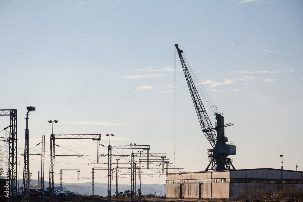 Industrial landscape at sunst, with an electrified railway line and its tracks, next to a heavy loading crane, standing next to a warehouse