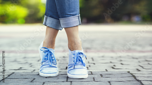 legs of a girl in jeans and blue sneakers on a sidewalk tile  a young woman strolling in a summer park