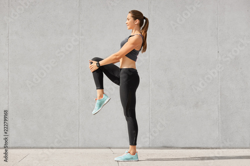 Shot of flexible young woman warms up before jogging on stadium, wears sportsclothes, shows her stomach and abs, poses against grey concrete wall. People, sport, fitness and flexibility concept