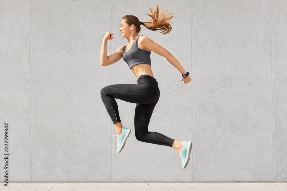 Active woman being full of energy, jumps high in air, wears sportsclothes, prepares for sport competitions, isolated over grey concrete wall. Female trainer busy with training. Gymnastics concept