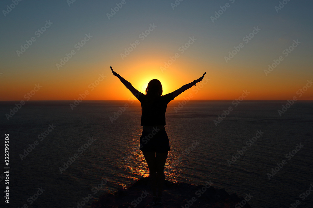 young girl is facing the sun on the mountain and looking at the sea, hands raised up and to the side
