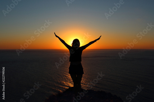 young girl is facing the sun on the mountain and looking at the sea  hands raised up and to the side
