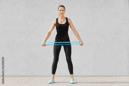Full length shot of attractive sporty woman dressed in black sportsclothes, holds rubber resistance band, has workout at home, poses against grey background. People, endurance, determination © sementsova321