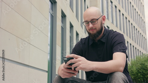 A man with a beard wearing black glasses uses a phone sitting in front of a large building. Medium shot. Soft focus © mputsylo