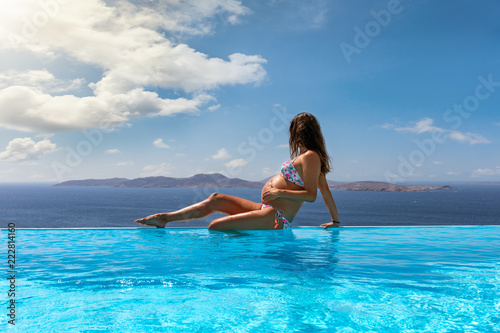 Schwangere Frau sitzt am Rande eines Infinity Pools und genießt die Aussicht auf das blaue Meer