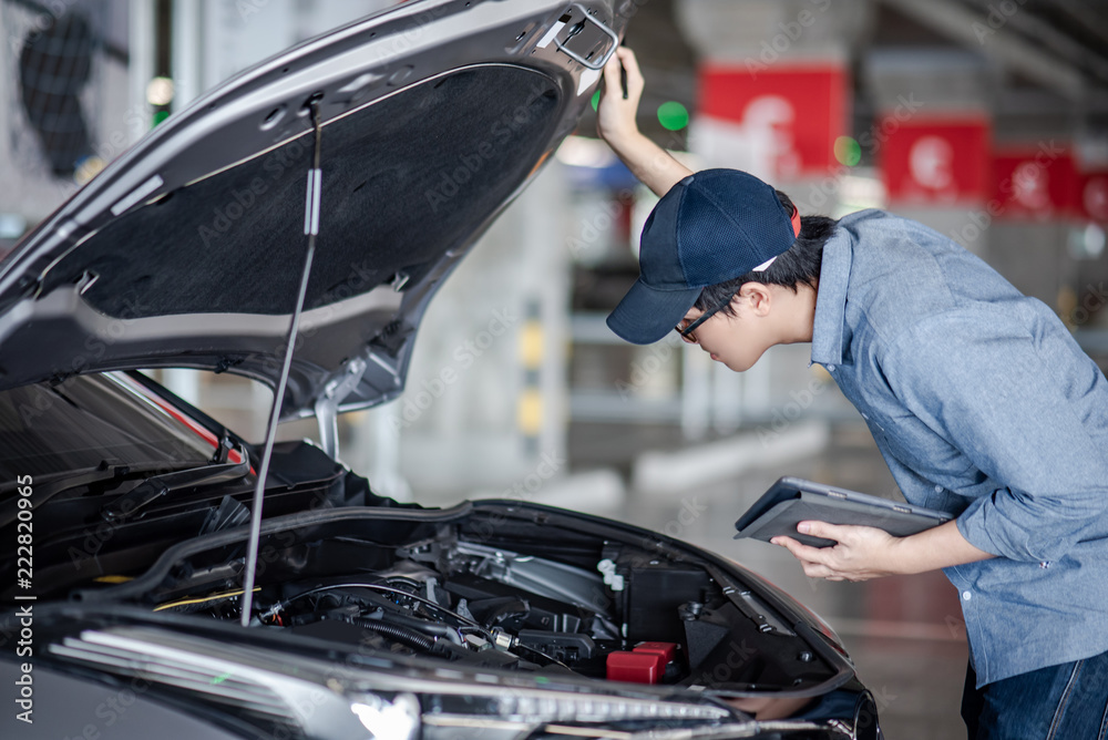 Obraz premium Asian auto mechanic holding digital tablet checking car engine under the hood in auto service garage. Mechanical maintenance engineer working in automotive industry. Automobile servicing and repair